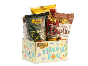 Thank you light yellow basket box with color full swirls/shapes, plants, and butterfliesgoing around the box with thank you written in blue with different colorful shapes on the letters and the unique snacks logo in the upper lefthand corner, filled with various Unique Snacks products